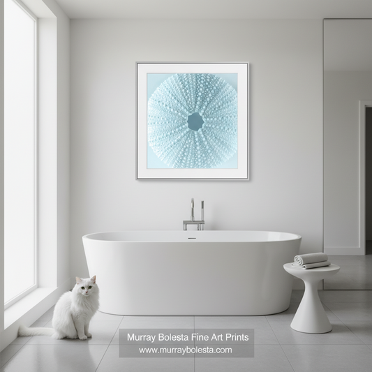 Blue sea urching by Murray Bolesta! A framed square art print featuring a light blue and white radial dot pattern hangs above a white freestanding bathtub in a bright modern bathroom. A white cat sits on the floor near the tub, and a small white side table holds folded towels.