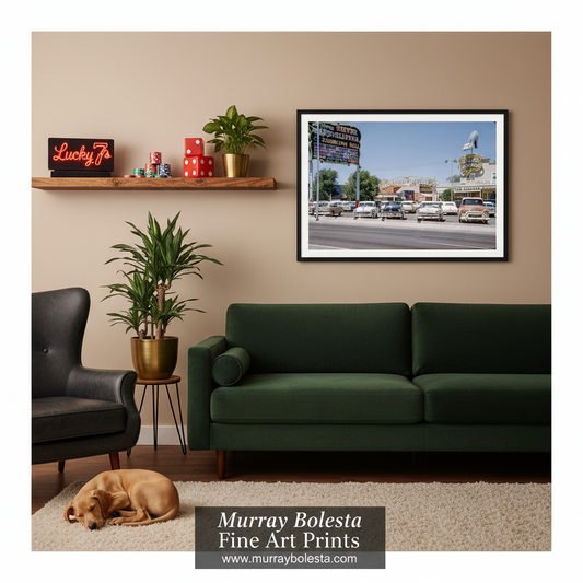 Las Vegas vintage. A framed fine art print of a street scene with vintage cars and a sign, displayed on a beige wall in a styled living room above a dark green sofa. The room includes a wooden shelf with decor, a potted plant, a black armchair, a sleeping dog on a rug, and a watermark at the bottom reading 'Murray Bolesta Fine Art Prints'.