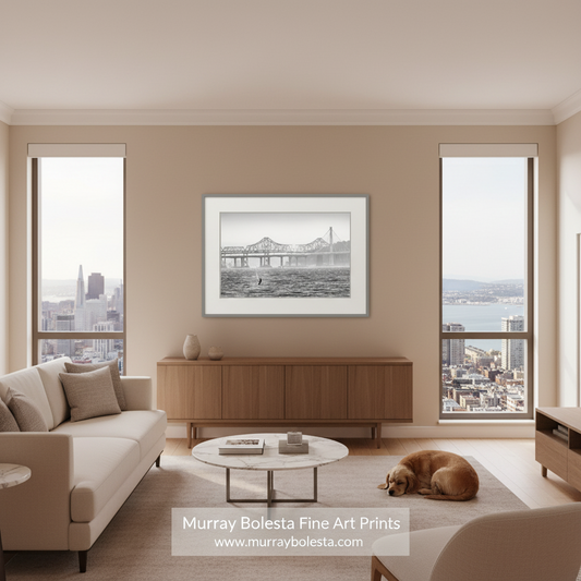 A framed black and white fine art print of a bridge over water hangs on a beige wall in a modern living room with a sofa, coffee table, and a sleeping dog on the floor. A watermark at the bottom reads 'Murray Bolesta Fine Art Prints'.