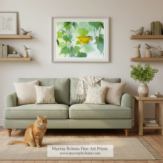 A framed art print featuring a yellow bird among green leaves, displayed on a beige wall above a sage green sofa in a styled living room. An orange tabby cat sits on the rug in the foreground.