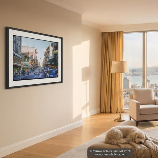A framed art print depicting a San Francisco city street scene with the Transamerica Tower, hung on a beige wall in a modern, sunlit living room with armchairs and a large window view. Mid-century road trip art by Murray Bolesta.