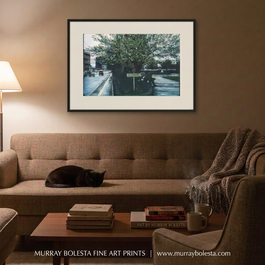 A framed art print of a street scene with a large tree, displayed on a beige wall above a brown sofa in a cozy living room setting with a sleeping black cat and a throw blanket. Georgia mid-century road trip art by Murray Bolesta