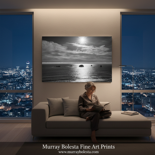 A black and white ocean landscape art print hangs on a beige wall above a gray sofa in a modern living room. A woman sits on the sofa reading, with a city skyline visible through large windows behind her. By Murray Bolesta.