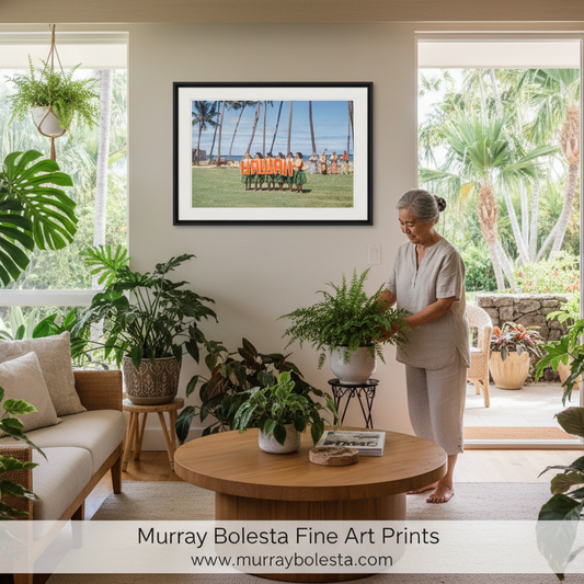 A framed art print of a beach scene with palm trees and a person sitting on a beach, displayed on a wall in a bright, modern living room with plants and a woman standing nearby. Midcentury hawaii road trip art by Murray Bolesta