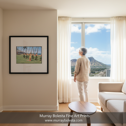 A framed art print depicting a Hawaiian hula dancer on a grassy field is displayed on a beige wall in a modern living room. An elderly woman stands by a large window looking out at a scenic mountain view under a blue sky. by murray bolesta.