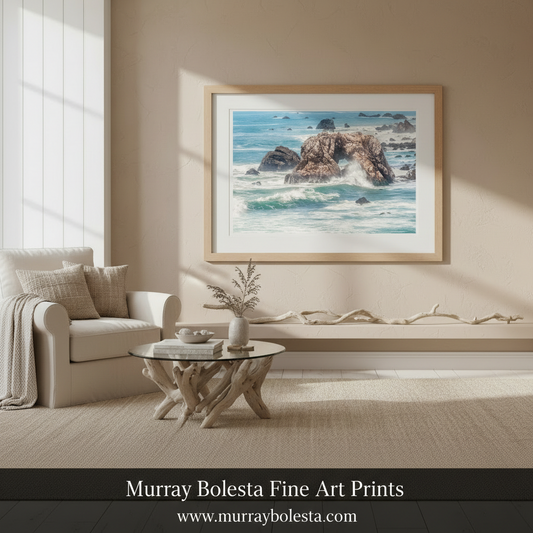 A framed coastal landscape art print hangs on a beige wall in a styled living room with a cream armchair, glass coffee table, and natural light streaming in from a window. By Murray Bolesta