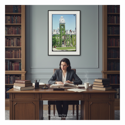 A framed art print depicting a green historic college building hangs on a gray wall above a wooden desk with a woman working in a study. The scene is framed by tall bookshelves on both sides. WVU by Murray Bolesta