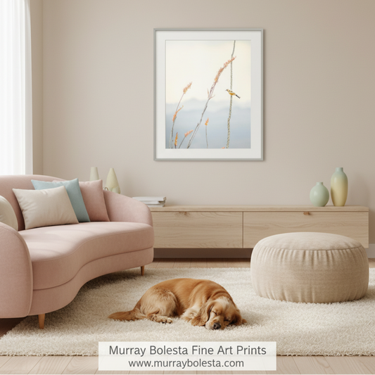 A framed art print featuring a minimalist landscape with a small bird and reeds, displayed on a beige wall in a styled living room above a light wood console, with a pink sofa, beige pouf, and a golden retriever dog lying on a white rug. A watermark at the bottom reads 'Murray Bolesta Fine Art Prints'. Bird art print!