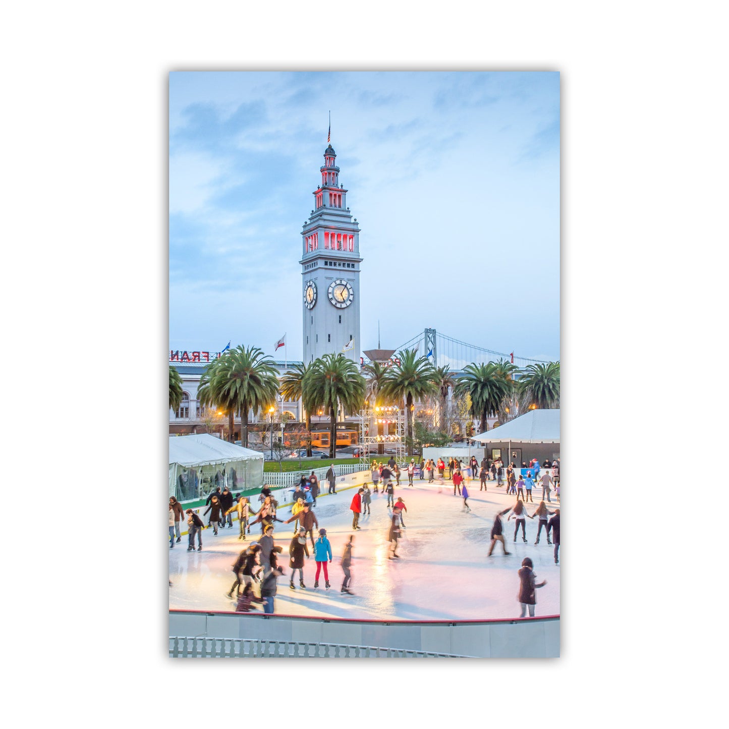 Ice skating rink with a clock tower and palm trees in the background. Beautiful Embarcadero! My splendid San Francisco photograph at Christmastime for your walls: twirling skaters in a holiday scene near the Ferry Building at the Embarcadero, with the Bay Bridge as a backdrop. Original, unique signed art by Murray Bolesta.