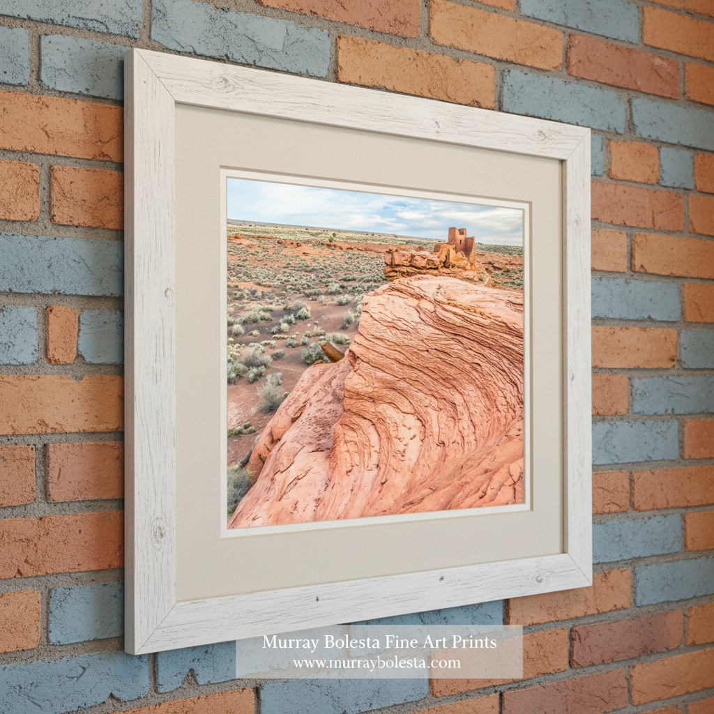 Desert landscape with red rock formations and a small building in the distance. Wupatki Monument by Murray Bolesta