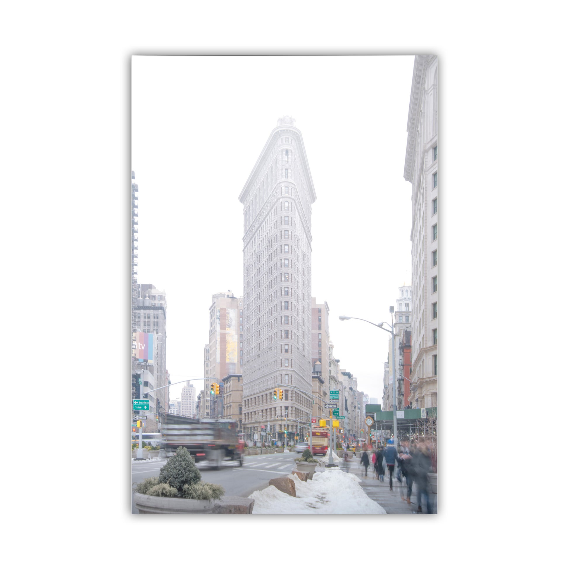 City street scene with a tall building in the center, framed by a white border. Flatiron Building NYC Winter Photo Print – Iconic Manhattan Landmark