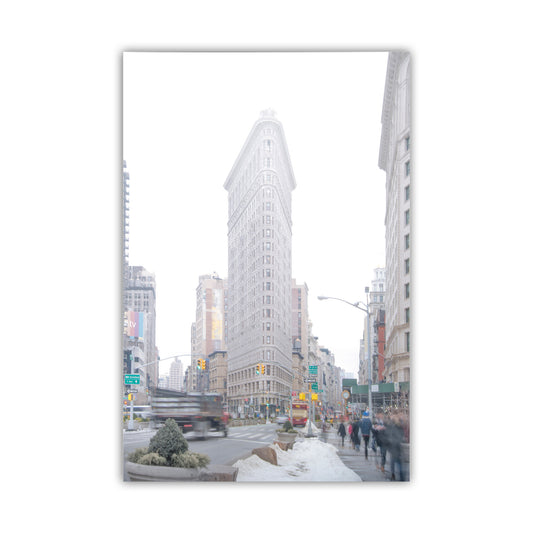 City street scene with a tall building in the center, framed by a white border. Flatiron Building NYC Winter Photo Print – Iconic Manhattan Landmark
