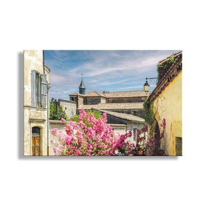 a picturesque European village scene, featuring colorful buildings, a church with a tall spire, and a vibrant flowering bush in the foreground. Quaint French Village Art Print | Vintage France Wall Decor