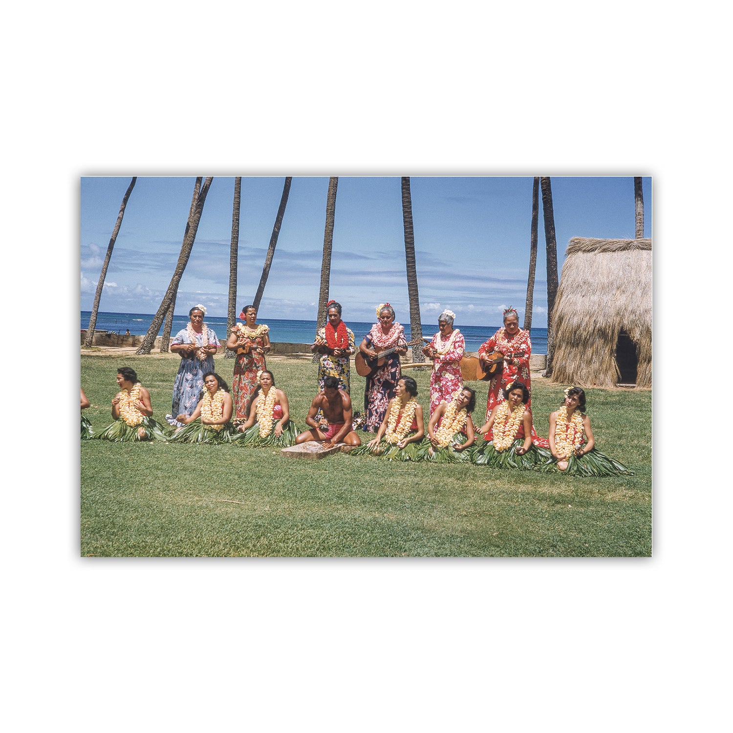 Group of people in traditional attire sitting on grass with palm trees and ocean in the background. Old Hawaii Photo of Hawaiian Islands Hula Dancers & Musicians