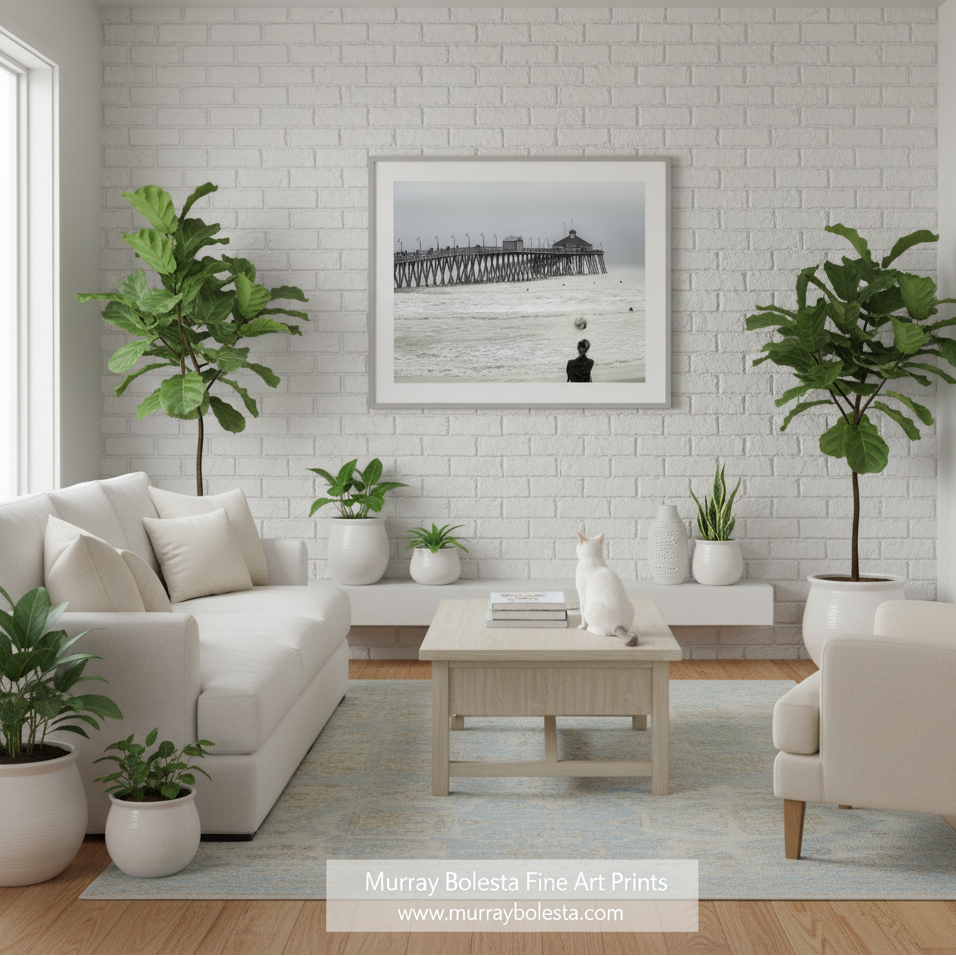 Imperial Beach San Diego! A framed black and white art print of a pier over the ocean hangs on a white brick wall in a bright, modern living room with a white sofa, plants, and a coffee table. A watermark at the bottom reads 'Murray Bolesta Fine Art Prints'.