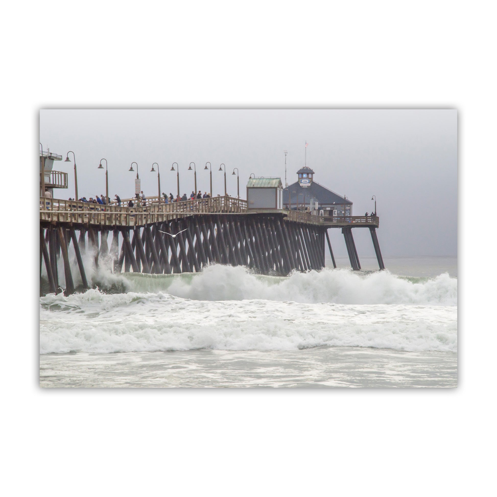 Pier with people on it surrounded by large waves on a cloudy day. Imperial Beach Pier Photo – Pacific Ocean Waves San Diego California