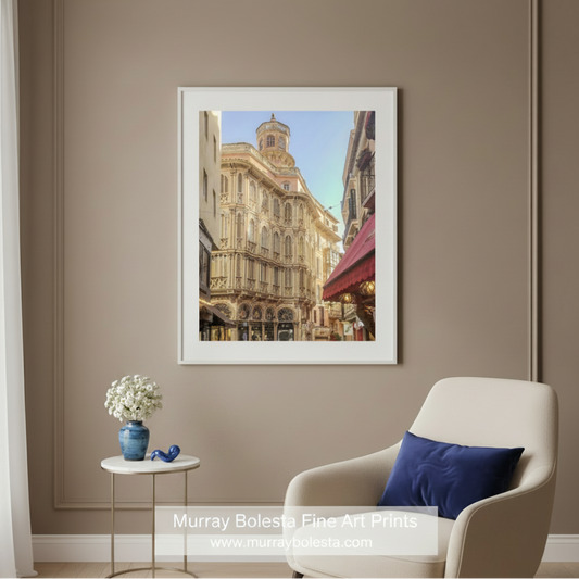 A framed art print of a European street scene with ornate beige buildings and a blue sky, displayed on a taupe wall above a cream armchair with a blue pillow and a small side table with flowers. Mallorca by Murray Bolesta