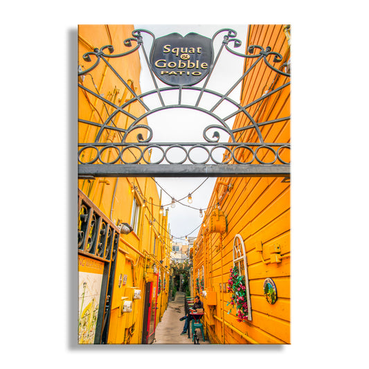 Colorful alleyway with yellow walls and a decorative metal archway labeled 'Squat & Gobble Patio'. San Francisco Street Photography Urban Art Print | Marina District
