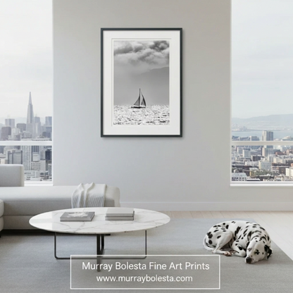 A framed black and white fine art print of a sailboat on water, displayed on a light gray wall in a modern living room with a gray sectional sofa, white coffee table, and a Dalmatian sleeping on the rug. The print is branded with 'Murray Bolesta Fine Art Prints' at the bottom. Sailing art San Francisco Bay!