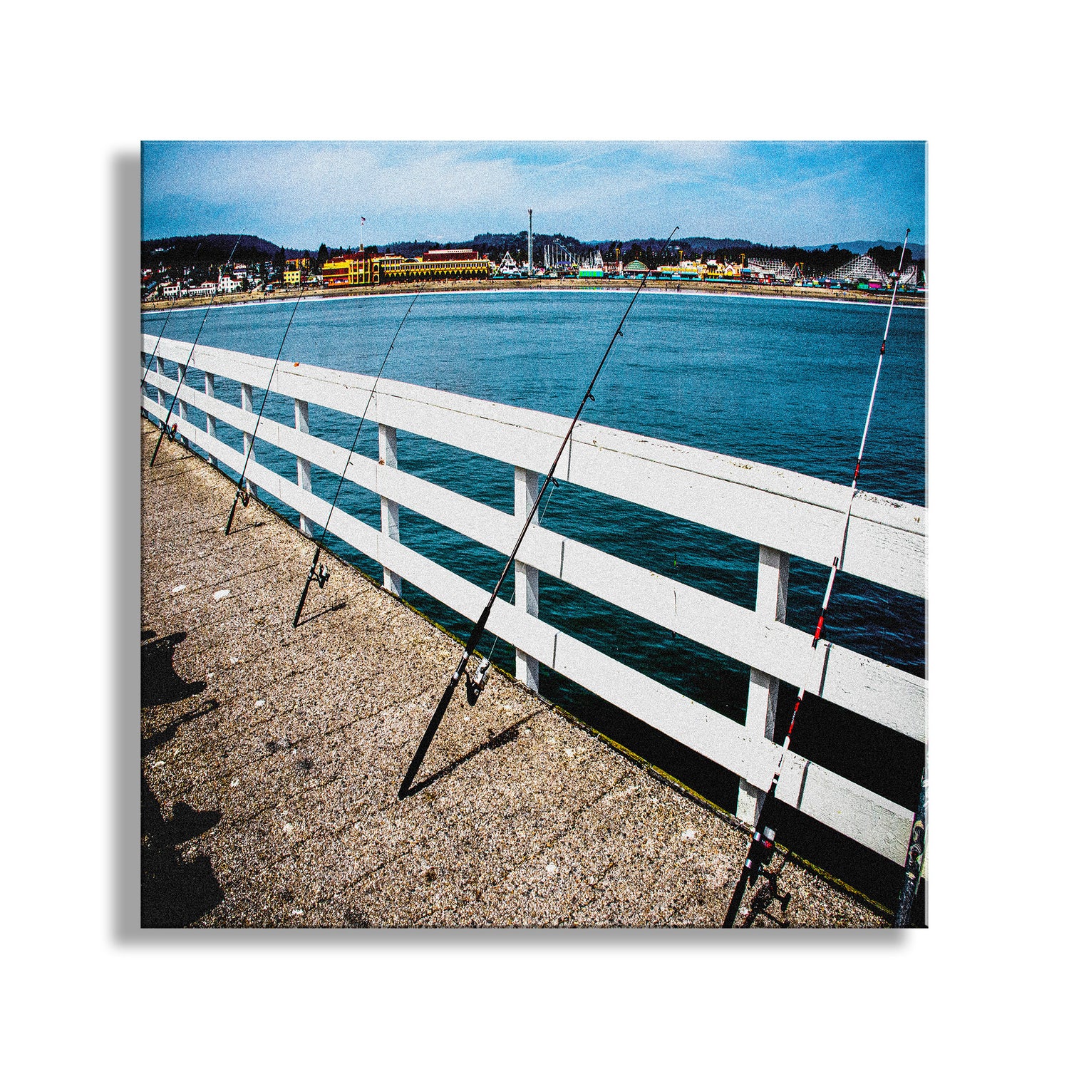 Fishing rods leaning against a white wooden railing by a body of water with a town in the background. Santa Cruz Wharf Picture with Beach Boardwalk on Monterey Bay