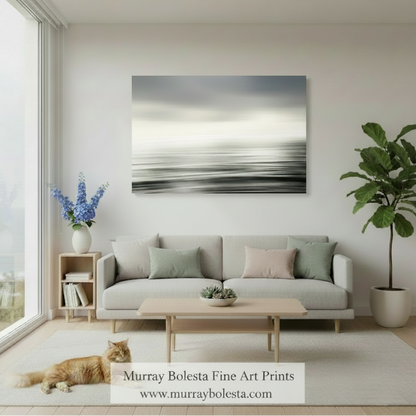 A framed abstract wall art print with soft gray, white, and black horizontal brushstrokes hangs above a light gray sofa in a bright, modern living room. An orange cat lies on the rug near a wooden coffee table, and a potted plant stands to the right.