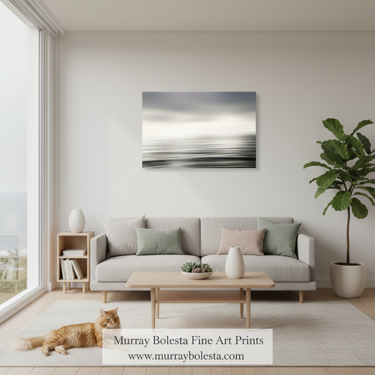 A framed abstract wall art print with soft gray, white, and black horizontal brushstrokes hangs above a light gray sofa in a bright, modern living room. An orange cat lies on the rug near a wooden coffee table, and a potted plant stands to the right.
