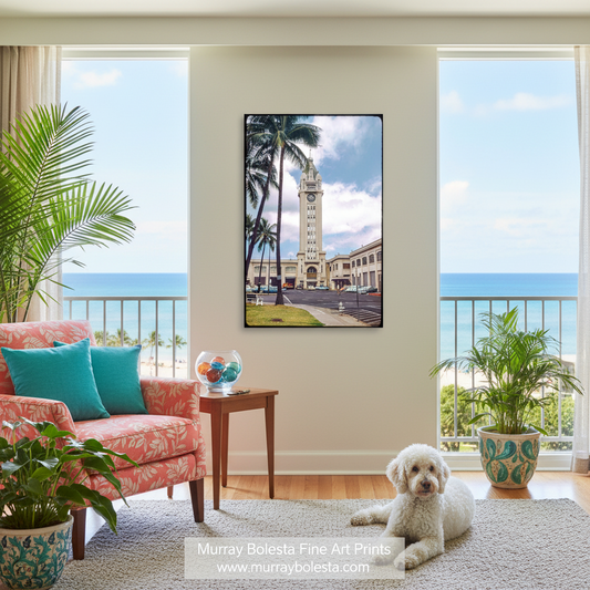 A framed vertical art print depicting a tropical coastal scene with palm trees, a clock tower, and the ocean, displayed on a wall in a bright living room with a pink armchair, plants, and a white dog.