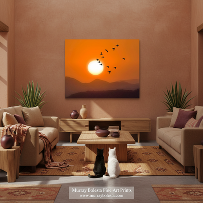 Arizona sunset art by Murray Bolesta! A framed wall art print showing an orange sunset over dark mountain silhouettes with flying birds, displayed above a coffee table in a styled living room with two cats sitting on the floor. 