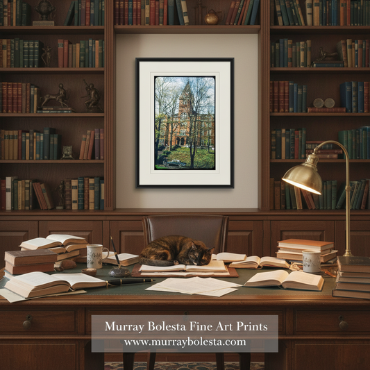 A framed art print depicting a historic campus building with trees is displayed on a wall in a detailed study setting. The scene includes a wooden desk cluttered with books, papers, mugs, and a gold desk lamp, with a cat sleeping in the chair.