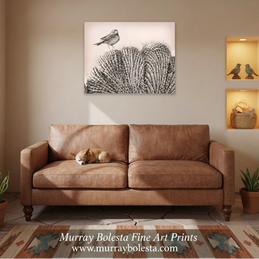 A square black and white fine art print of a bird perched on a textured fan of feathers, displayed on a beige wall above a brown leather sofa in a styled living room. Bird art by Murray Bolesta