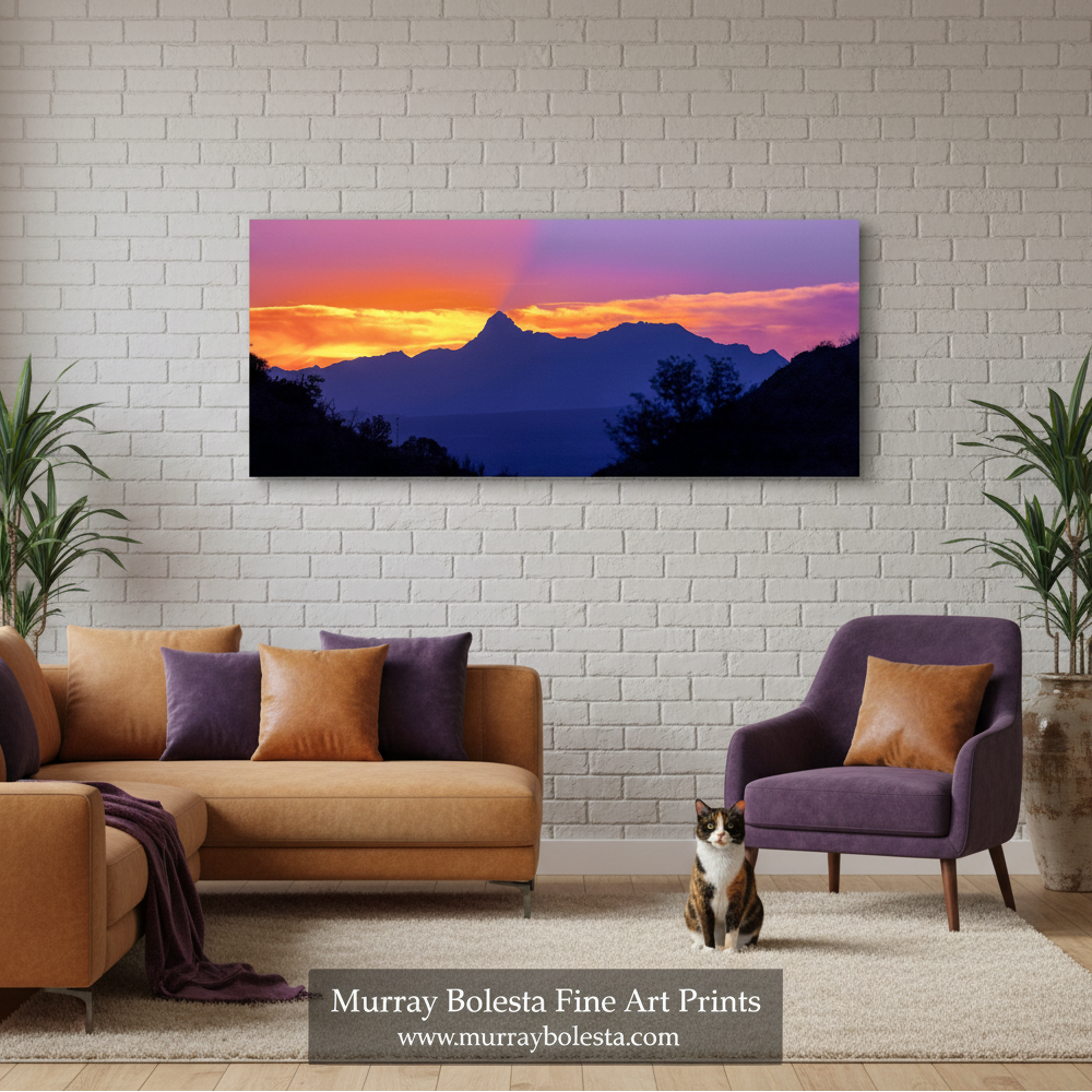 Baboquivari Peak Arizona by Murray Bolesta. A rectangular wall art print showing a mountain sunset with purple, orange, and pink sky, displayed above a sofa in a modern living room. The print is the product being sold, and the room setting is a styled interior mockup.