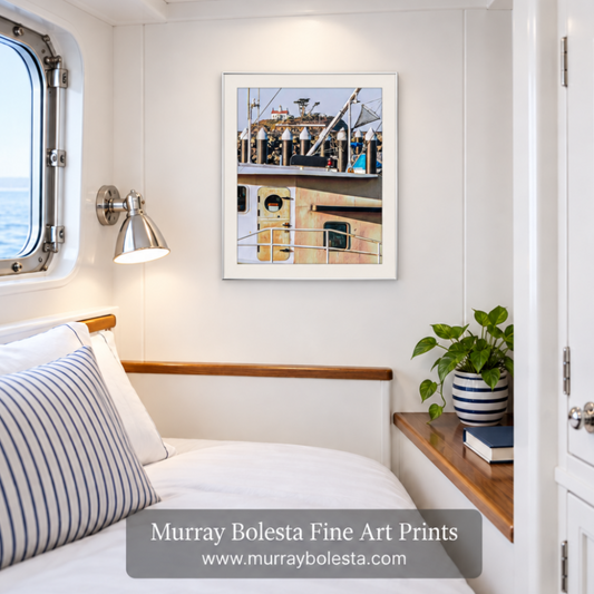 A framed art print of a fishing boat deck hanging on a white wall in a nautical cabin setting. The print features a white frame and is displayed above a bed with striped pillows and next to a wooden shelf with a potted plant. Battery point lighthouse by Murray Bolesta