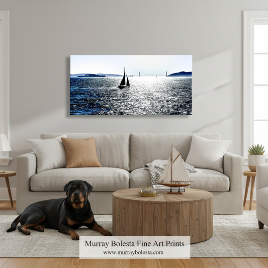 A framed canvas art print depicting a sailboat on a blue ocean under a pale sky, displayed on a light gray wall above a beige sofa in a modern living room. A black and tan dog lies on the rug in front of the couch, and a wooden coffee table with a model sailboat sits in the foreground.