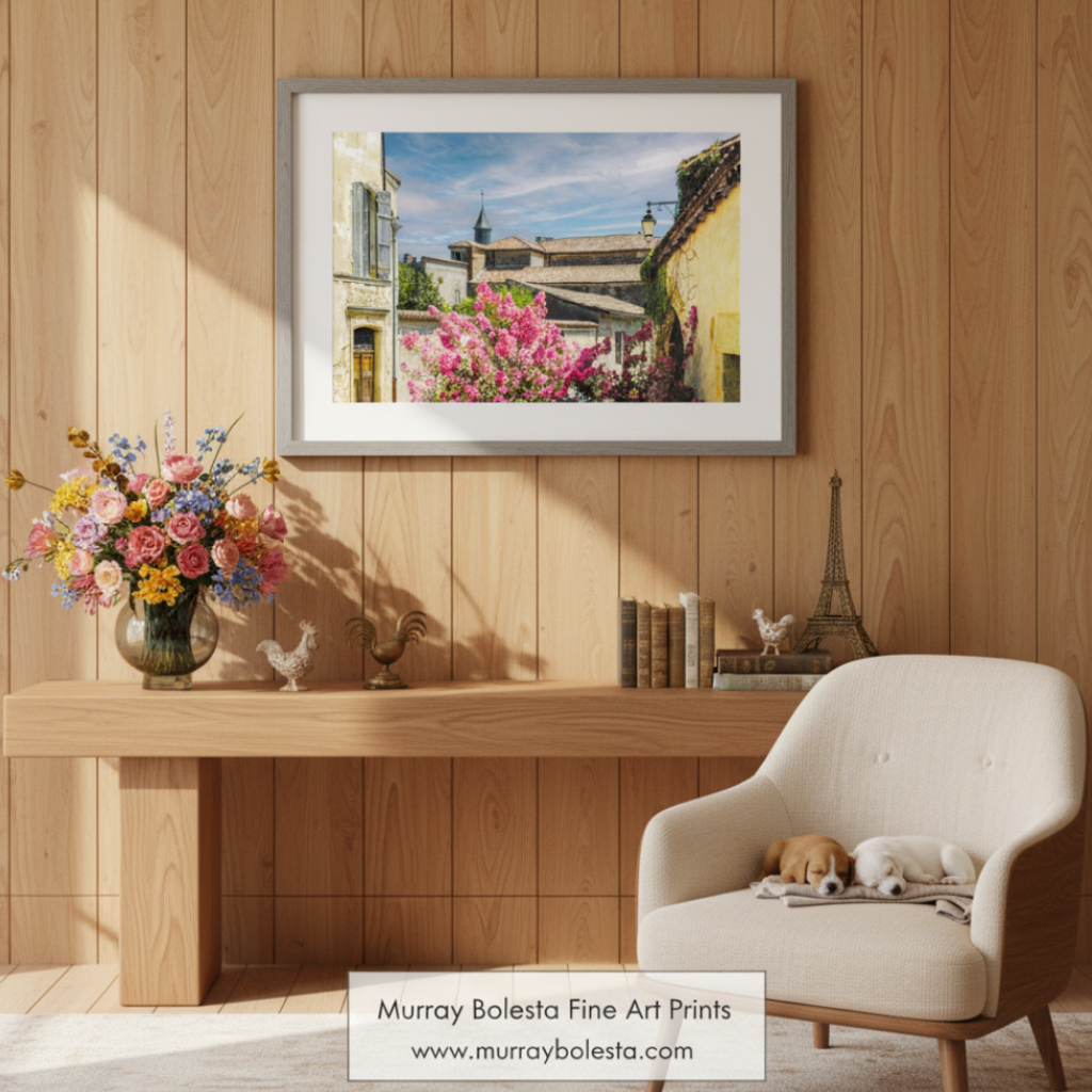 a picturesque European village scene, featuring colorful buildings, a church with a tall spire, and a vibrant flowering bush in the foreground. Quaint French Village Art Print | Vintage France Wall Decor by Murray Bolesta