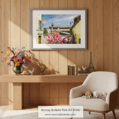 a picturesque European village scene, featuring colorful buildings, a church with a tall spire, and a vibrant flowering bush in the foreground. Quaint French Village Art Print | Vintage France Wall Decor by Murray Bolesta