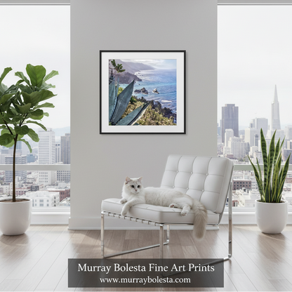 Big Sur agave by Murray Bolesta. A framed art print of a coastal landscape with cacti and ocean waves is displayed on a light gray wall in a modern living room setting. A white cat is lying on a white tufted chair in the foreground, with a city skyline visible through the large windows behind.