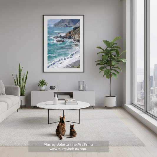 Big Sur California Pacific Coast Highway by Murray Bolesta! A framed vertical art print of a coastal ocean scene with rocky cliffs and turquoise waves, displayed on a light gray wall in a modern living room. Two cats sit on a white rug below the print, with a round coffee table, plants, and large windows nearby.