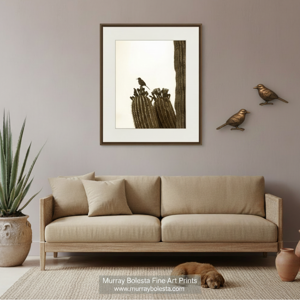 A framed art print featuring a saguaro cactus with birds, displayed on a gray wall above a beige fabric sofa in a styled living room setting. Desert bird art by Murray Bolesta!
