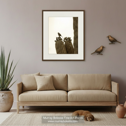 A framed art print featuring a saguaro cactus with birds, displayed on a gray wall above a beige fabric sofa in a styled living room setting. Desert bird art by Murray Bolesta!