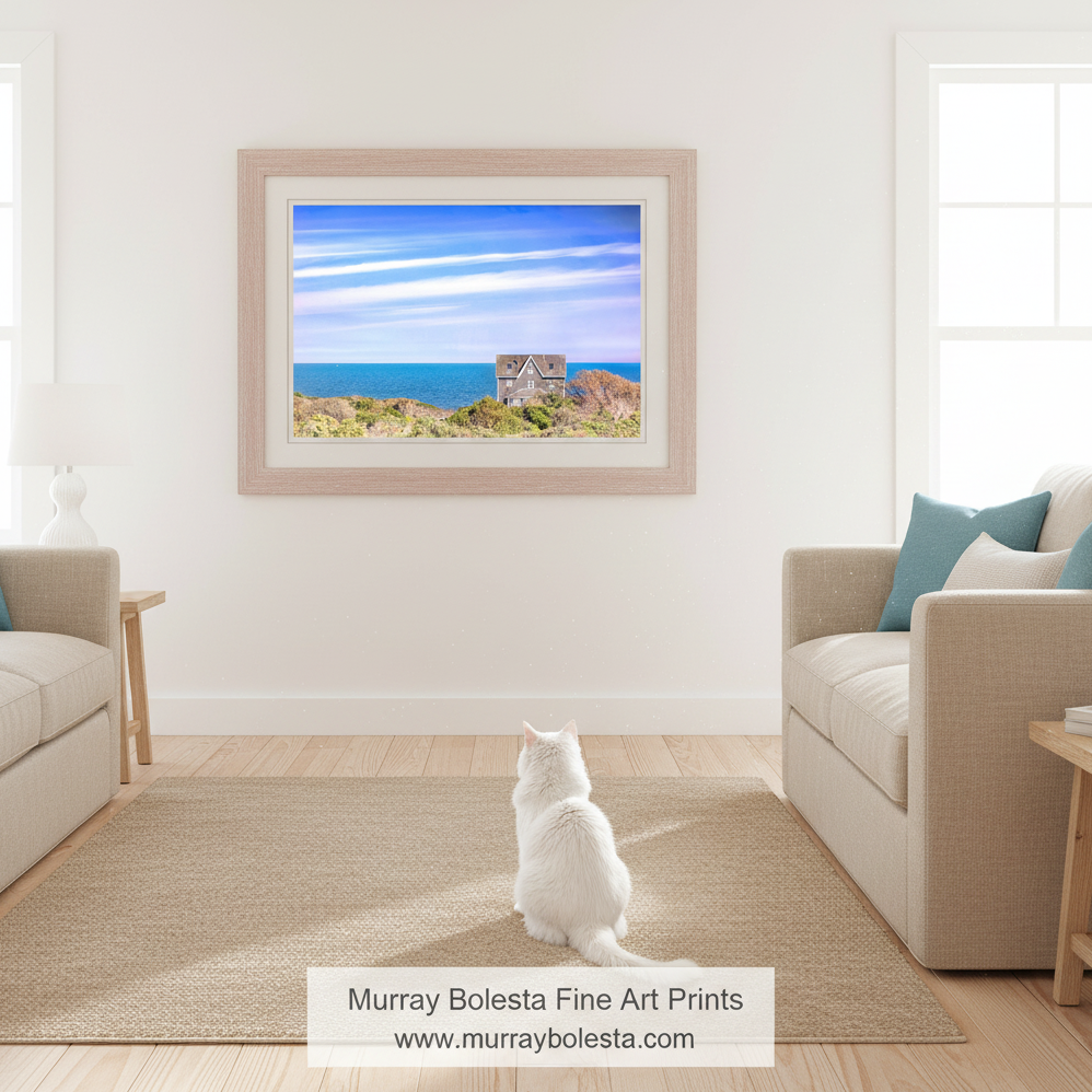A two-story house with a slanted roof sits on a cliff overlooking the ocean, surrounded by lush greenery and a clear blue sky with wispy clouds. Block Island Seaside Cottage Art - Rhode Island Coastal Wall Decor by Murray Bolesta