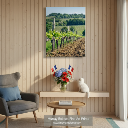 A row of grapevines with green leaves and small white flowers, surrounded by a dirt path and a church spire in the distance. France Vineyard Picture Bordeaux | Wine Country Landscape Art by Murray Bolesta