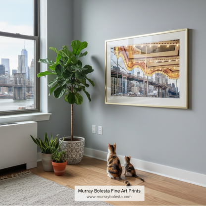 A framed art print depicting a New York City bridge and skyline hangs on a gray wall in a modern room. Two cats are sitting on the wooden floor below, looking at the print. Janes Carousel artwork by Murray Bolesta Fine Art Prints!