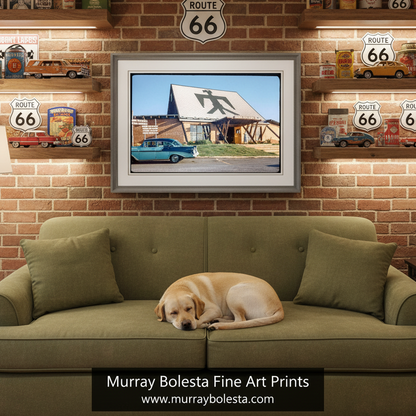 A framed art print depicting a Route 66 roadside scene with a blue car and a building, displayed on a brick wall above an olive green sofa with a dog resting on it. Decorative Route 66 signs and shelves are visible in the background. Vintage mid-century road trip art by Murray Bolesta