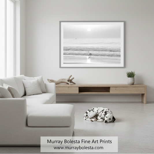 A framed black and white landscape art print hangs on a light gray wall above a wooden console in a modern, minimalist living room. A white sectional sofa is on the left, and a black-and-white dog is lying on the floor. Rhode Island Beach art by Murray Bolesta