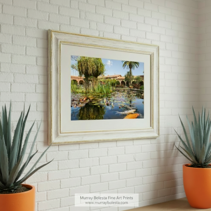 A tranquil pond with lily pads, surrounded by palm trees and a building with arched windows, creating a picturesque scene. San Juan Capistrano Mission Artwork - California Church Print by Murray Bolesta