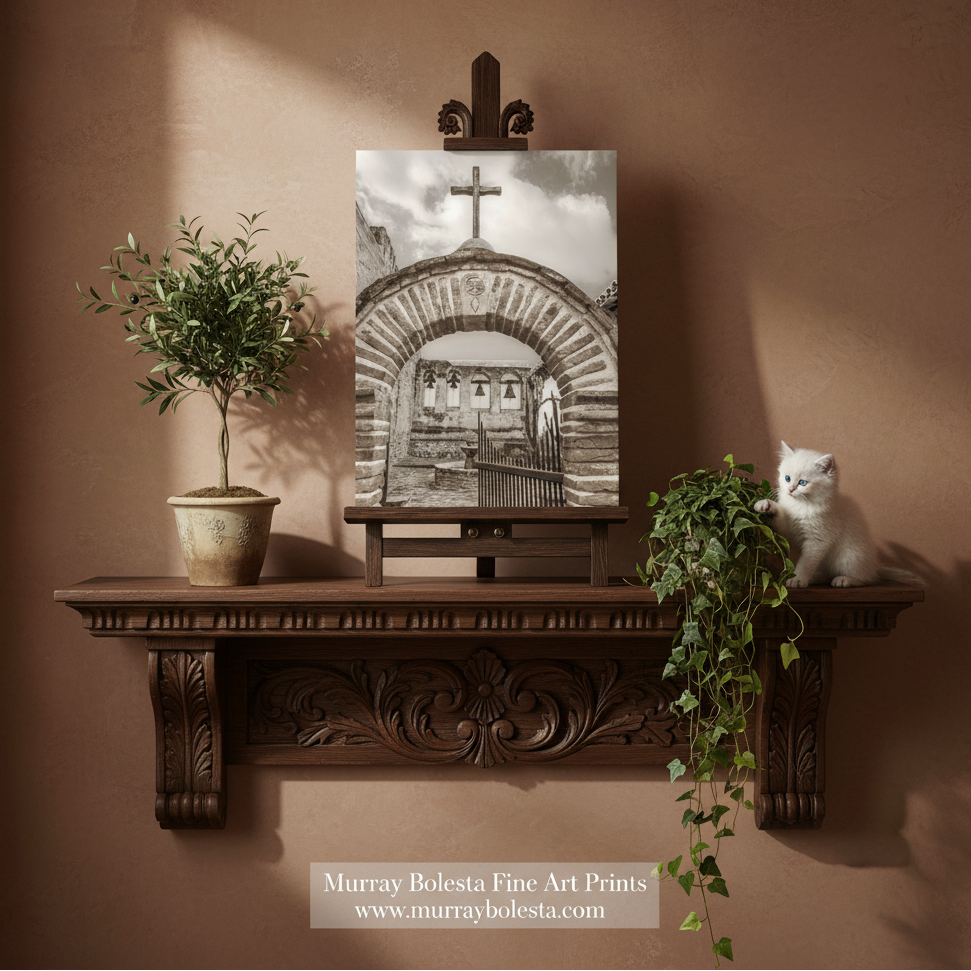 Capistrano Mission by Murray Bolesta. A sepia art print of a cathedral archway with a cross, displayed on a wooden easel on a dark brown carved mantelpiece. The scene is styled with a potted olive tree on the left and a white cat sitting on the mantel on the right, all against a textured warm brown wall.