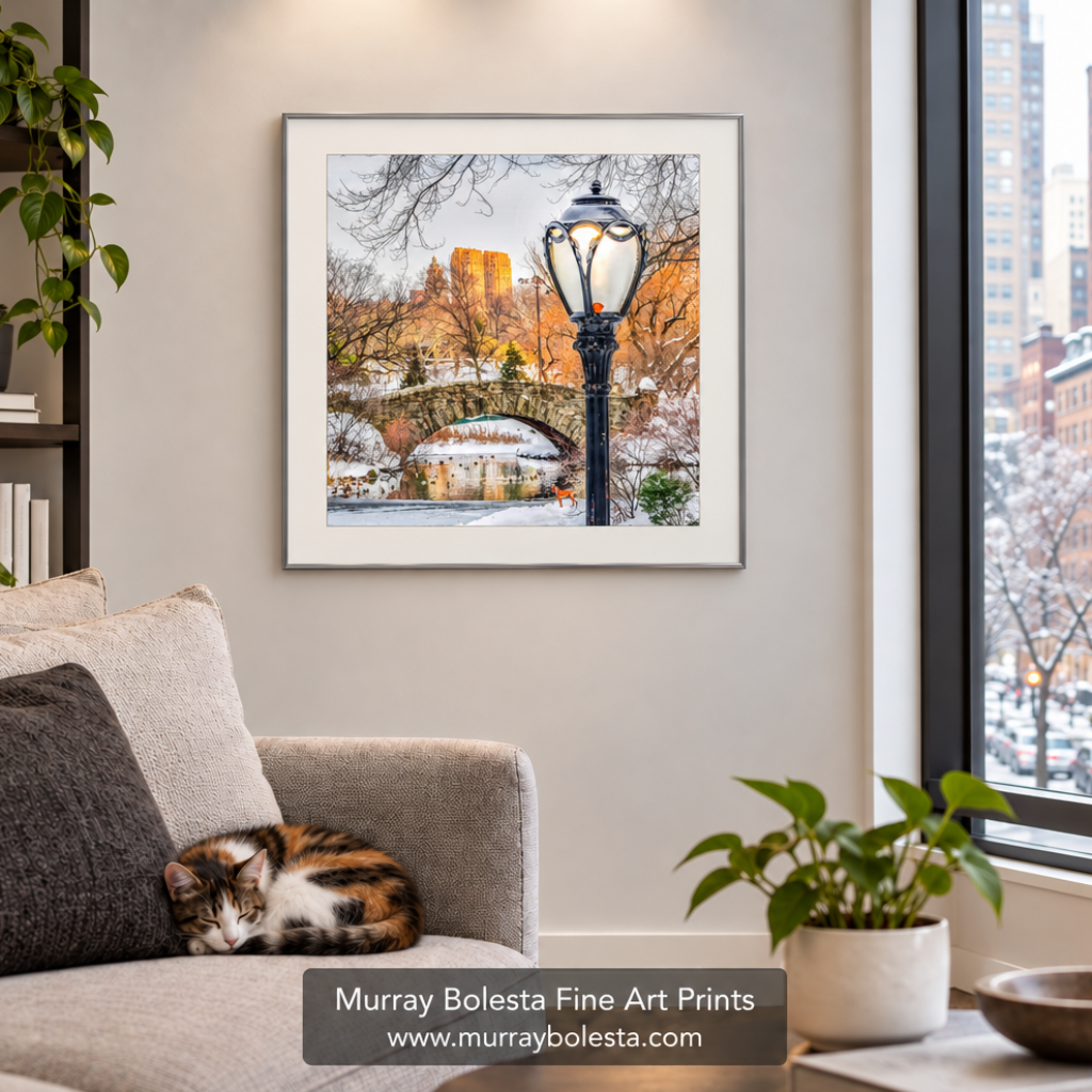 A framed art print of a snowy Central Park scene with a bridge, fountain, and a black lamppost, displayed on a beige wall above a gray sofa with a cat resting on it. A window on the right shows a city view, and a watermark for 'Murray Bolesta Fine Art Prints' is at the bottom. Art by Murray Bolesta.