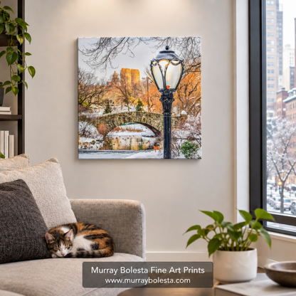 A square wall art print showing a snowy urban park scene with a bridge, street lamp, and warm-toned buildings, displayed above a sofa in a living room. A cat rests on the couch, and a window view of the city is visible. Central Park Winter art by Murray Bolesta.