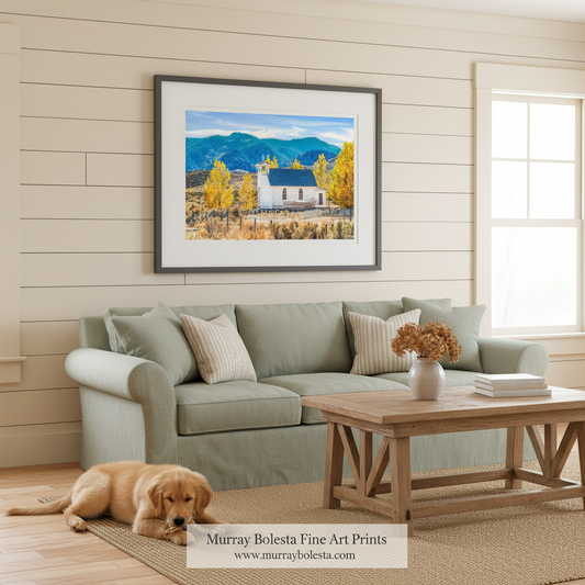 A framed landscape art print of a white church with yellow autumn trees and blue mountains, displayed on a beige shiplap wall above a sage green sofa with a golden retriever lying on the floor.