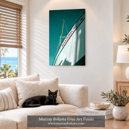 A vertical canvas art print featuring a teal and white sailboat design with the owall in a styled living room. A black cat lies on a cream-colored sofa in the foreground, and a window on the left shows a view of the ocean and palm trees. Sailing art by Murray Bolesta
